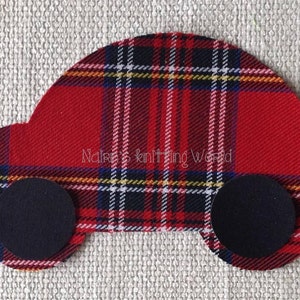 Op de afbeelding: Een stoffen applicatie in de vorm van een auto, met een rood, zwart, wit en geel ruitpatroon en twee effen zwarte ronde wielen. De tekst "Naira's knitting World" is in een lichte kleur gedrukt over het midden van de autovorm.