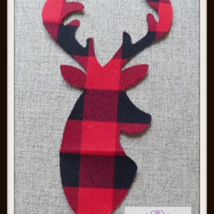 May include: A fabric deer head silhouette featuring a red and black buffalo plaid pattern. The deer has prominent antlers and pointed ears. A rectangular label in the bottom right corner displays the purple text Nairncraft with decorative swirls.