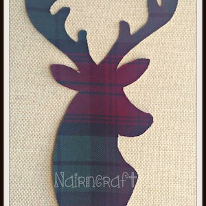 Può includere: Una sagoma in tessuto a forma di testa di cervo con corna, caratterizzata da un motivo tartan a quadri rosso scuro, verde e blu. La parola Nairncraft è stampata in lettere bianche stilizzate sulla parte inferiore della sagoma del cervo.