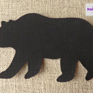 May include: A black fabric bear silhouette applique with a textured surface. The bear is depicted in profile facing left with a visible snout and four legs. In the top right corner, there is a white rectangular label with the text Nairncraft.