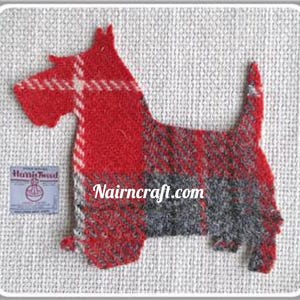May include: A Scottish Terrier shaped fabric applique made of red and grey plaid Harris Tweed wool. The design features a textured weave with a white cross-hatch pattern. A small rectangular Harris Tweed label is attached, and the text Nairncraft.com is printed in white across the center of the dog figure.