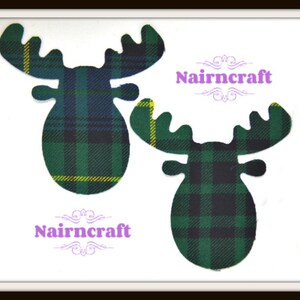 May include: Two fabric moose head cutouts featuring a green, black, and yellow plaid tartan pattern. The word Nairncraft is printed in purple text with decorative flourishes above and below the name, appearing twice on the white background.