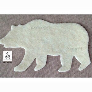 May include: A cream-colored felt bear cutout facing left. The bear has a textured surface and a soft appearance. A small white square label with a black orb and cross logo and the text Harris Tweed Left Facing is visible in the lower left corner.