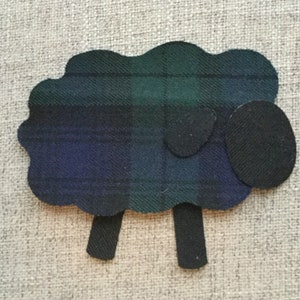 May include: A sheep-shaped fabric appliqué featuring a body made of dark green, blue, and black plaid tartan fabric. The head, two legs, and one ear are crafted from solid black felt material, creating a distinct contrast against the patterned body.
