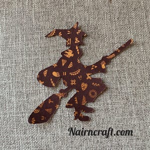Könnte beinhalten: Ein dunkelbrauner Stoffzuschnitt in Form einer Hexe auf einem Besen mit einem wiederkehrenden Muster aus kleinen orangefarbenen Symbolen wie Herzen, Sternen und Pfeilen. Der Text Nairncraft.com ist unten rechts in Schwarz aufgedruckt.