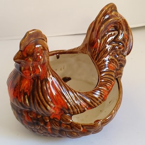 May include: A ceramic hen-shaped planter with a brown and orange glaze. The planter has a large opening for plants and is designed with detailed feathers and a rooster's comb. The interior is a cream color.
