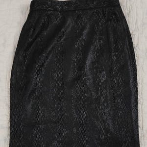 May include: A black knee-length skirt featuring a textured, snakeskin-like pattern throughout the fabric. The garment has a structured waistband and a straight, tailored silhouette that maintains a consistent width from the waist to the hem.
