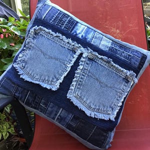 K&ouml;nnte beinhalten: Ein quadratisches Dekokissen aus verschiedenen blauen Jeansstoffen. Das Design zeigt zwei markante Jeans-Taschen mit ausgefransten, rohen Kanten, die in der Mitte aufgen&auml;ht sind. Die Oberfl&auml;che zeigt ein Patchwork aus verschiedenen Jeans-Texturen.