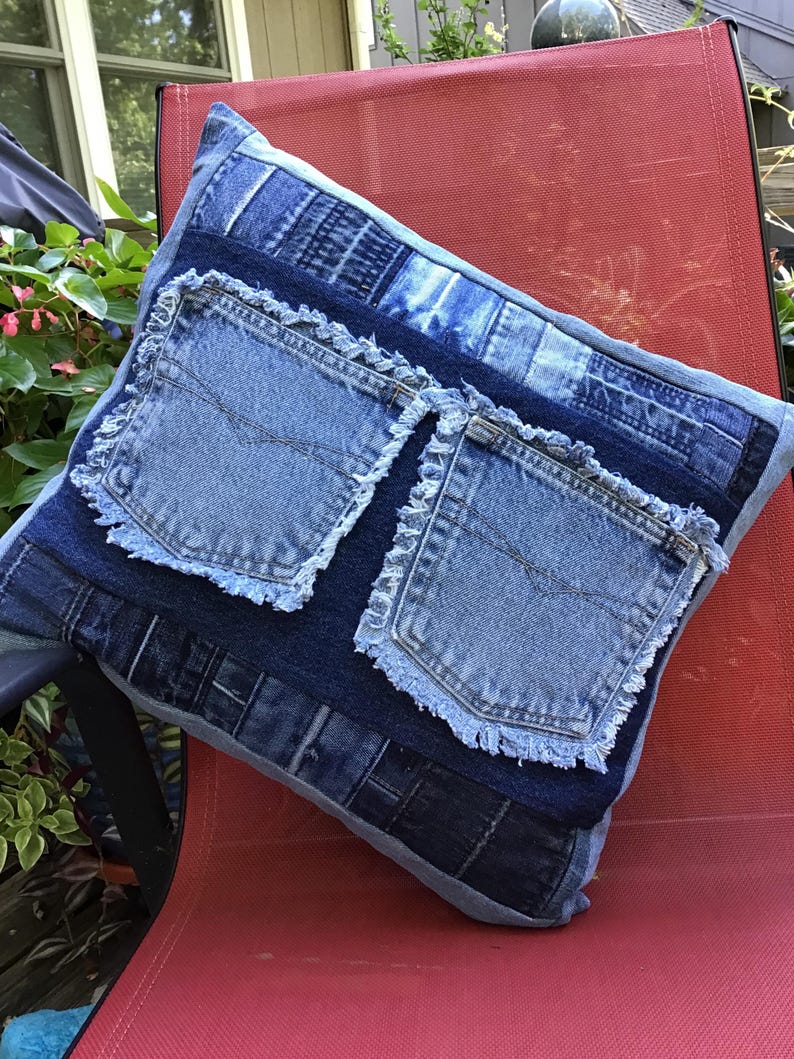K&ouml;nnte beinhalten: Ein quadratisches Dekokissen aus verschiedenen blauen Jeansstoffen. Das Design zeigt zwei markante Jeans-Taschen mit ausgefransten, rohen Kanten, die in der Mitte aufgen&auml;ht sind. Die Oberfl&auml;che zeigt ein Patchwork aus verschiedenen Jeans-Texturen.