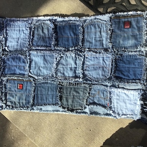 May include: A rectangular denim rug made from repurposed jeans. The rug features a patchwork design of various blue jean pockets and denim squares, with a fringed edge. Some pockets have red tags.