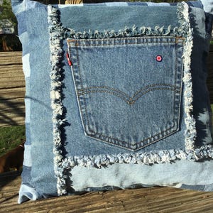 May include: Square denim pillow with a patchwork design. The pillow features a light blue denim pocket with a red tag and a small pink and blue emblem. The edges are trimmed with frayed denim.