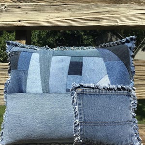 May include: A square throw pillow constructed from various shades of blue denim fabric patches. The surface features a patchwork design with a prominent denim pocket sewn onto the front, and all edges are finished with a frayed, raw denim fringe.