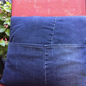 K&ouml;nnte beinhalten: Ein quadratisches Dekokissen aus dunkelblauem Jeansstoff. Das Kissen verf&uuml;gt &uuml;ber eine horizontale Naht in der Mitte und eine gro&szlig;e, funktionale Tasche, die sich &uuml;ber die gesamte Breite des unteren Bereichs erstreckt, mit sichtbaren N&auml;hten und Denim-Struktur.