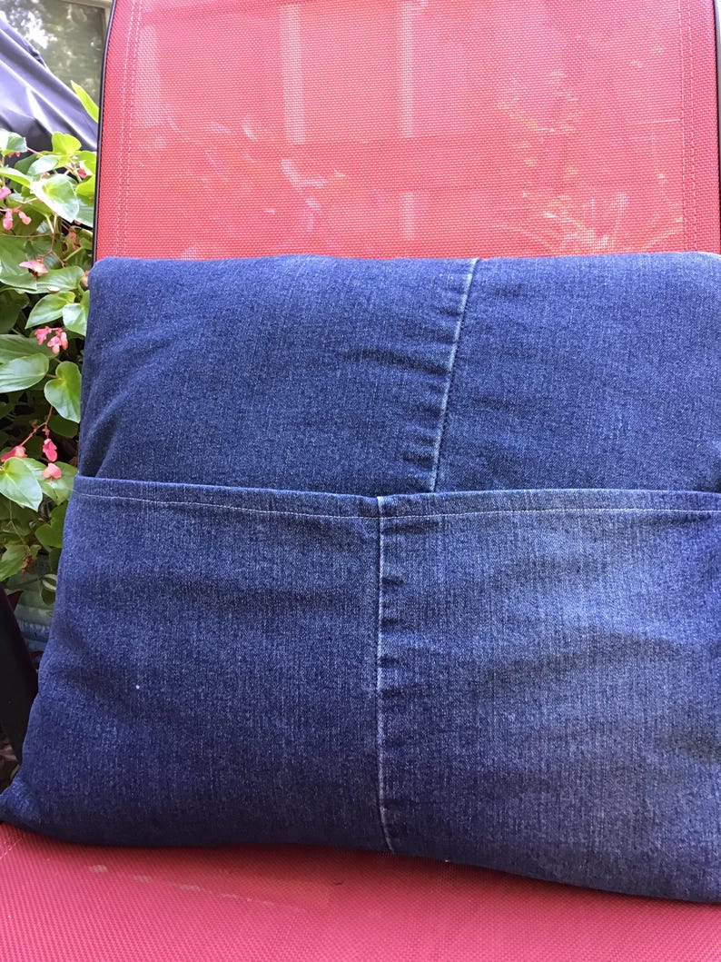 K&ouml;nnte beinhalten: Ein quadratisches Dekokissen aus dunkelblauem Jeansstoff. Das Kissen verf&uuml;gt &uuml;ber eine horizontale Naht in der Mitte und eine gro&szlig;e, funktionale Tasche, die sich &uuml;ber die gesamte Breite des unteren Bereichs erstreckt, mit sichtbaren N&auml;hten und Denim-Struktur.