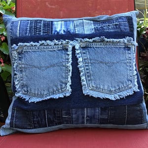 K&ouml;nnte beinhalten: Ein quadratisches Zierkissen aus einem Patchwork verschiedener blauer Jeansstoffe. In der Mitte befinden sich zwei dunkelblaue Jeanstaschen mit ausgefransten Kanten. Die umliegenden Bereiche bestehen aus verschiedenen Blaut&ouml;nen von Jeansstreifen.