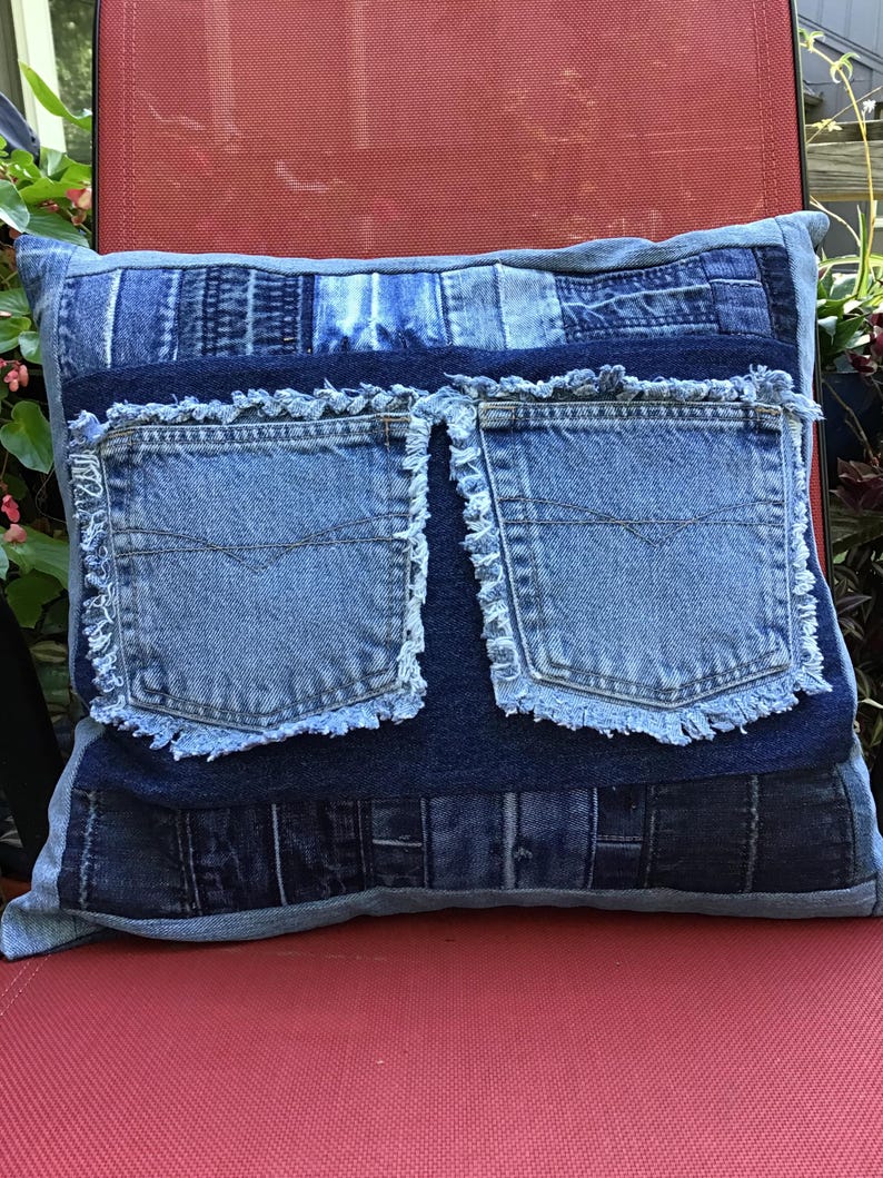 K&ouml;nnte beinhalten: Ein quadratisches Zierkissen aus einem Patchwork verschiedener blauer Jeansstoffe. In der Mitte befinden sich zwei dunkelblaue Jeanstaschen mit ausgefransten Kanten. Die umliegenden Bereiche bestehen aus verschiedenen Blaut&ouml;nen von Jeansstreifen.