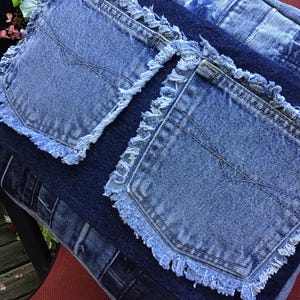K&ouml;nnte beinhalten: Ein quadratisches Zierkissen aus wiederverwertetem blauem Jeansstoff. Die Oberfl&auml;che zeigt ein Patchwork-Design aus verschiedenen Blaut&ouml;nen, akzentuiert durch zwei aufgesetzte Jeans-Taschen mit ausgefransten R&auml;ndern auf der Vorderseite.