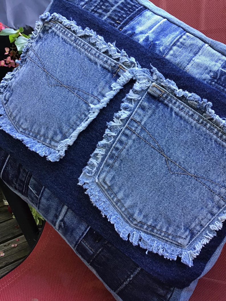 K&ouml;nnte beinhalten: Ein quadratisches Zierkissen aus wiederverwertetem blauem Jeansstoff. Die Oberfl&auml;che zeigt ein Patchwork-Design aus verschiedenen Blaut&ouml;nen, akzentuiert durch zwei aufgesetzte Jeans-Taschen mit ausgefransten R&auml;ndern auf der Vorderseite.