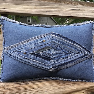May include: A rectangular denim pillow with a diamond-shaped design made from repurposed denim. The pillow has a frayed edge and is in shades of blue. The diamond design is embellished with small brass-colored studs.
