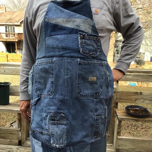 May include: A denim apron with multiple pockets and a patchwork design. The apron is made from recycled blue jeans and features a small pocket on the chest. The straps are a blue and white checkered pattern.