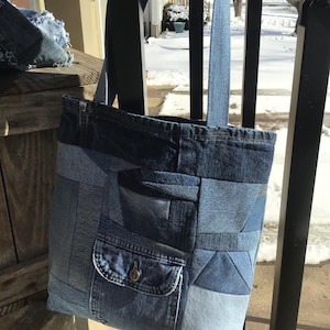 May include: A patchwork denim tote bag with a shoulder strap. The bag features a variety of blue denim patches, including a pocket with a button closure. The bag is hanging on a black metal railing.