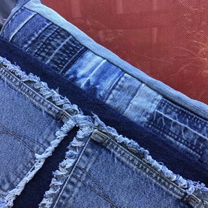 K&ouml;nnte beinhalten: Ein rechteckiges Zierkissen aus verschiedenen Blaut&ouml;nen von Jeansstoff. Die Oberfl&auml;che weist ein Patchwork-Design mit sichtbaren ausgefransten Kanten, freiliegenden N&auml;hten und mehreren auf die Vorderseite aufgen&auml;hten Jeanstaschen auf.