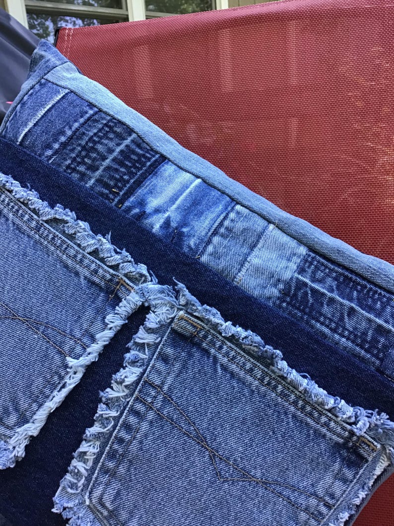 K&ouml;nnte beinhalten: Ein rechteckiges Zierkissen aus verschiedenen Blaut&ouml;nen von Jeansstoff. Die Oberfl&auml;che weist ein Patchwork-Design mit sichtbaren ausgefransten Kanten, freiliegenden N&auml;hten und mehreren auf die Vorderseite aufgen&auml;hten Jeanstaschen auf.