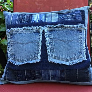 May include: A square throw pillow constructed from a patchwork of various blue denim fabrics. The center features a dark blue denim panel with two attached light blue denim pockets, each displaying frayed, raw edges along their rectangular borders.