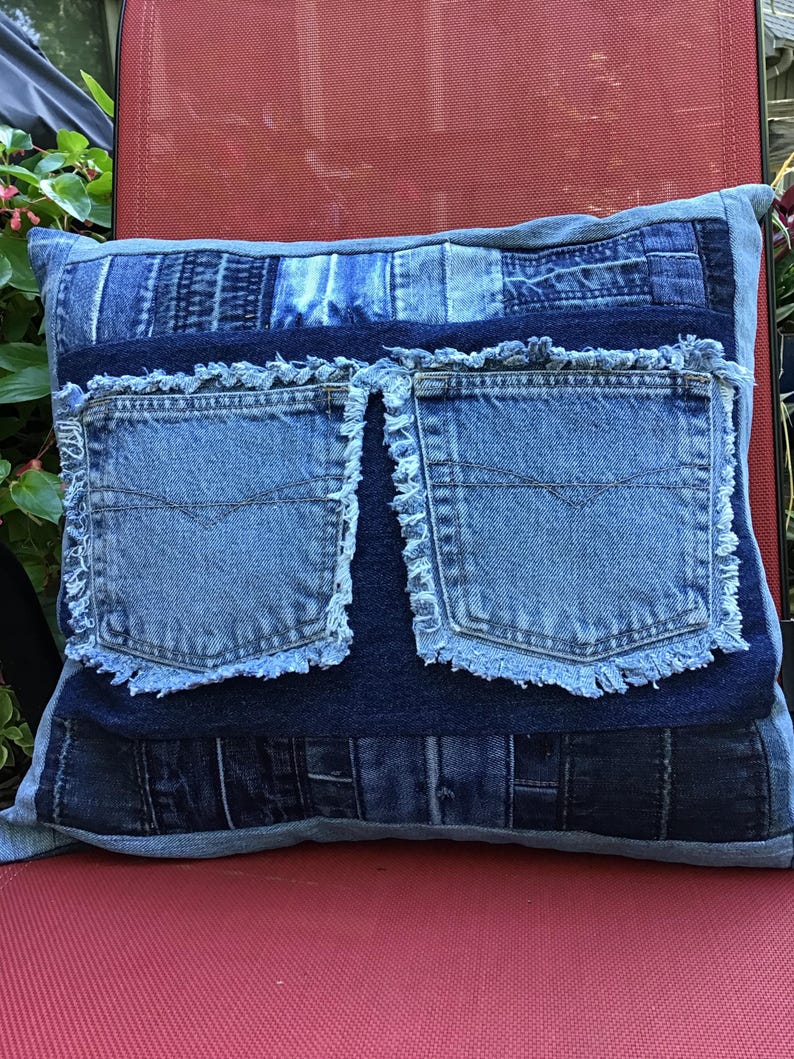 K&ouml;nnte beinhalten: Ein quadratisches Zierkissen aus einem Patchwork verschiedener blauer Jeansstoffe. In der Mitte befindet sich ein dunkelblaues Jeanspanel mit zwei aufgesetzten hellblauen Jeanstaschen, die jeweils ausgefranste Kanten an ihren R&auml;ndern aufweisen.