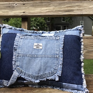 May include: A rectangular throw pillow constructed from various shades of blue denim fabric. The center features a light blue denim pocket with a white Lee Dungarees label. The edges of the pillow and the pocket are finished with frayed denim.