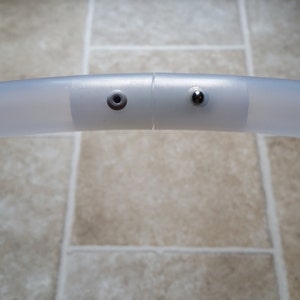 Puede incluir: Primer plano de un aro de pl&aacute;stico blanco con dos conectores de metal plateado. Los conectores tienen un peque&ntilde;o agujero redondo y un peque&ntilde;o bot&oacute;n redondo.