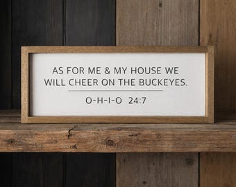 May include: A rectangular wooden sign with a white background and a light brown frame. The text reads: AS FOR ME & MY HOUSE WE WILL CHEER ON THE BUCKEYES. O-H-I-O 24:7. The sign features black lettering and a horizontal line separating the two phrases.