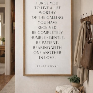 May include: A framed wall art print with a wooden frame displays the text "I URGE YOU TO LIVE A LIFE WORTHY OF THE CALLING YOU HAVE RECEIVED. BE COMPLETELY HUMBLE + GENTLE. BE PATIENT. BEARING WITH ONE ANOTHER IN LOVE. EPHESIANS 4:1". A woven basket and a coat rack are also visible.