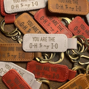 May include: A collection of leather keychains in various colors, including red, brown, and gray. Each keychain has a rectangular shape with rounded corners and a metal ring. The text "YOU ARE THE O-H to my I-O" is embossed on each keychain.