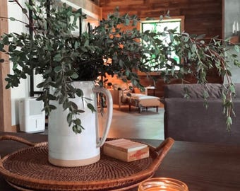 May include: A white ceramic pitcher with a tan base holds green leafy branches, resting on a woven tray. A small wooden box and a lit candle in a glass jar sit nearby. The scene is set on a dark wooden table.