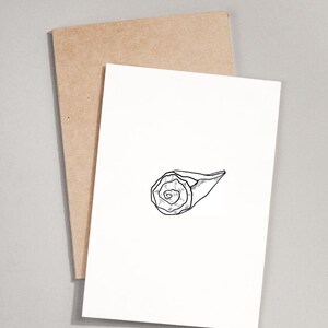 May include: A white greeting card featuring a black ink line drawing of a rolled-up cinnamon bun or pastry in the center. The card is positioned in front of a plain, light brown kraft paper envelope, creating a simple and clean visual presentation.