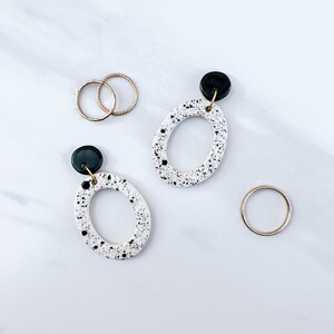 May include: A pair of white oval earrings with black speckles and dark green accents. The earrings are dangling from gold hooks.