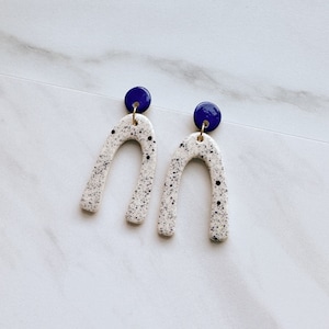 May include: A pair of white ceramic earrings with a speckled blue design. The earrings are shaped like an arch and have a small blue circle at the top.