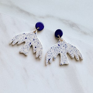 May include: A pair of white ceramic earrings with a blue splatter pattern in the shape of a bird. The earrings have a blue button-shaped accent at the top.