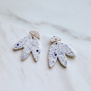 May include: A pair of white ceramic earrings with a blue speckled pattern. The earrings are shaped like a flower with three petals.