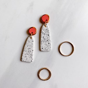 May include: A pair of white ceramic earrings with black speckles and a coral red top. The earrings are hanging from gold hoops. There are two gold rings on a white surface.