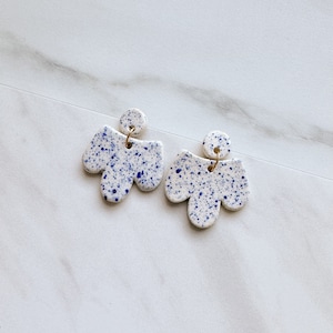 May include: A pair of white ceramic earrings with blue speckles. The earrings are shaped like a cluster of three rounded shapes.