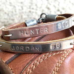 May include: Two personalized leather bracelets with silver nameplates. One reads "HUNTER #10" and the other "JORDAN #6". The bracelets are brown and rest on a brown leather surface, likely a baseball glove.