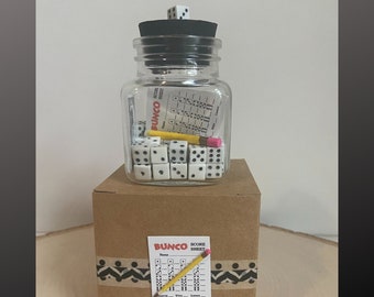 May include: A clear glass jar filled with white dice, a yellow pencil, and a score sheet. The jar is topped with a black lid and a single die. The jar sits on a brown cardboard box decorated with a decorative tape and a score sheet.