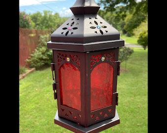May include: A decorative black metal lantern with red glass panels. The lantern has a geometric design with cut-out patterns and is adorned with colorful gems. The lantern is hanging outdoors with a green background.