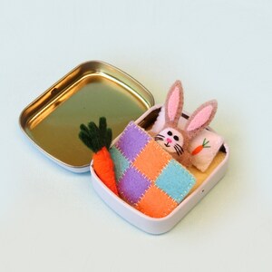 May include: A small, white tin with a gold interior, open to reveal a felt bunny with pink ears, a patchwork blanket, a carrot, and a pillow with a carrot design. The bunny is nestled inside.