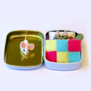 May include: A small white mouse with pink ears sits next to a tin box. The box contains a green cat with yellow and blue eyes sleeping in a patchwork quilt with pink, yellow, blue and green squares.