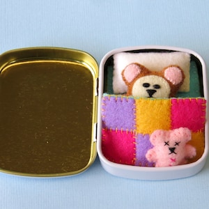 May include: A small metal tin containing a felt bear resting on a white pillow under a multicoloured patchwork quilt. A smaller pink felt bear with stitched facial features sits on top of the quilt. The tin has a gold-coloured interior base.