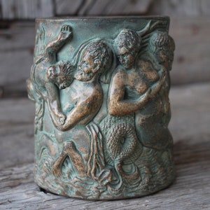 May include: A ceramic mug with a green and brown glaze features a raised design of a mythical creature with a human torso and a fish tail. The creature is holding a human figure in its arms.
