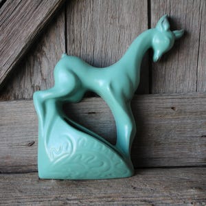 May include: A turquoise ceramic sculpture of a stylized deer with its head bent down. The deer is standing on a base with a decorative pattern.